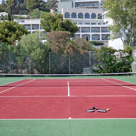 Hotel Dolce By Wyndham Athens Attica Riviera Vravrona Exterior foto The photo shows a tennis court with red and green surfaces. The court is empty, with no players present. In the center, there are a tennis racket and a ball lying on the ground. In the background, there are trees and a building, suggesting the court 
