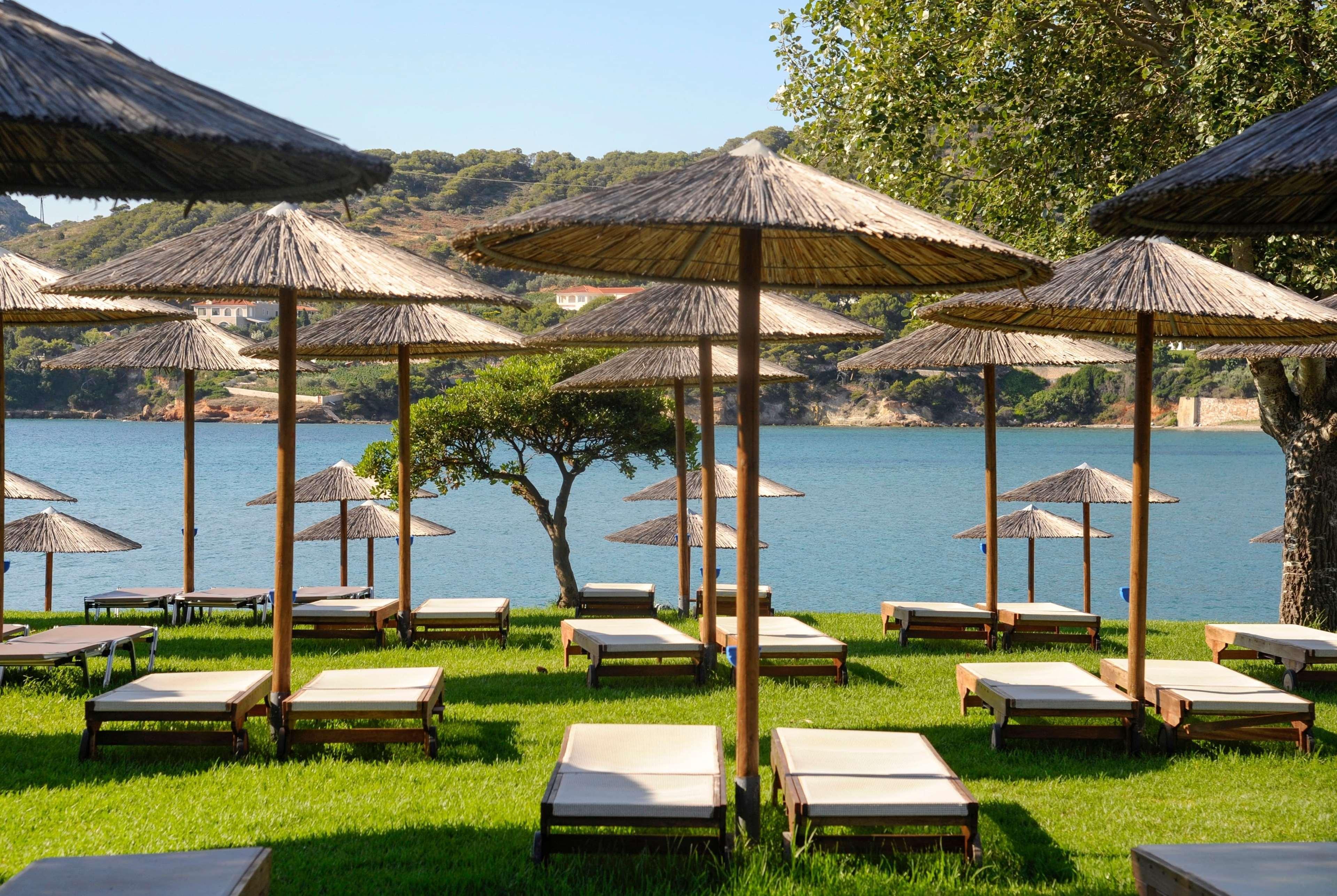 Hotel Dolce By Wyndham Athens Attica Riviera Vravrona Exterior foto The photo shows a serene beach scene with several lounge chairs arranged on a green lawn. Each lounge chair is shaded by a traditional straw umbrella, providing a relaxing spot to enjoy the view. In the background, there is a calm body of water, like