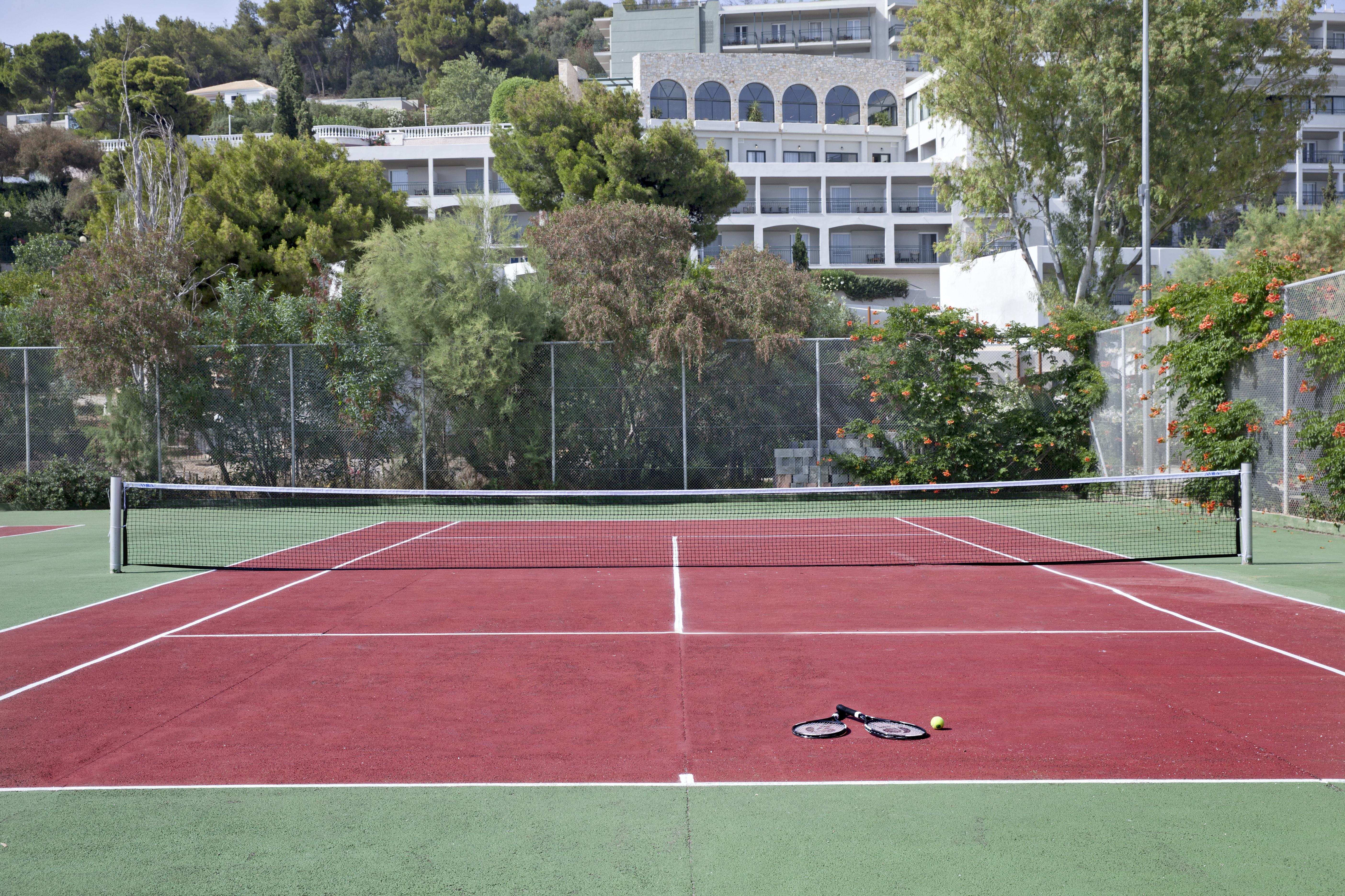 Hotel Dolce By Wyndham Athens Attica Riviera Vravrona Exterior foto The photo shows a tennis court with red and green surfaces. The court is empty, with no players present. In the center, there are a tennis racket and a ball lying on the ground. In the background, there are trees and a building, suggesting the court 