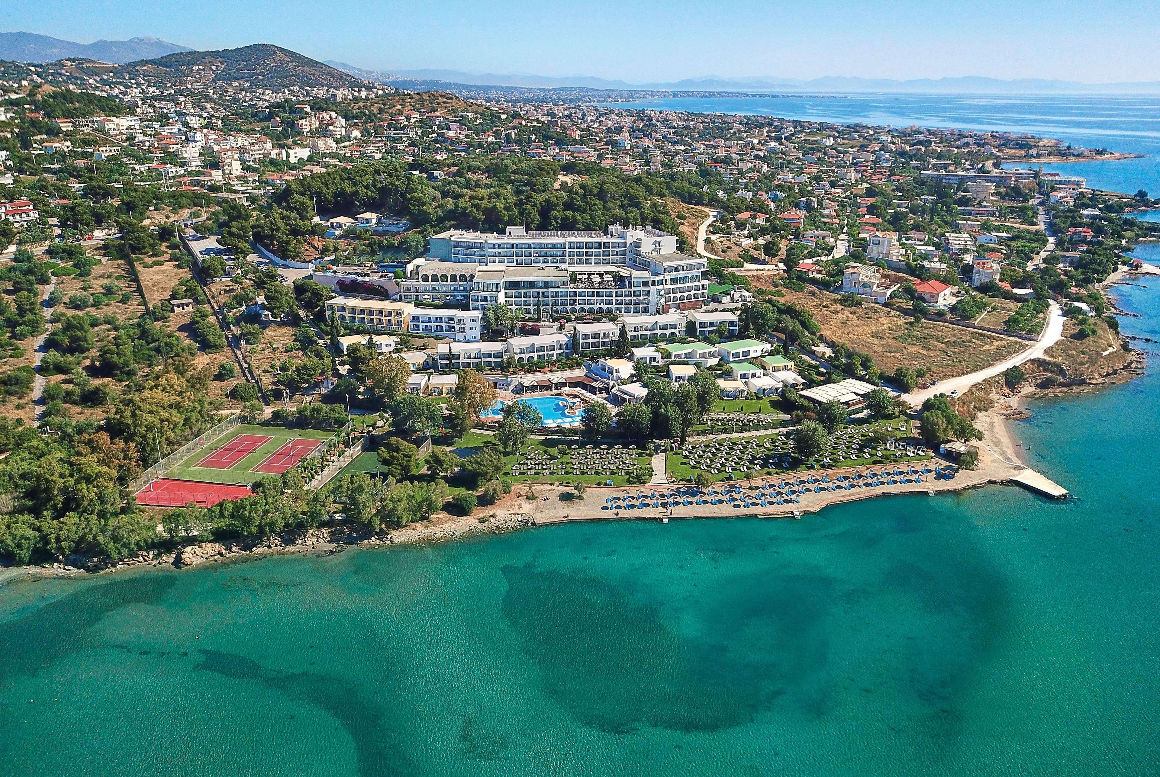 Hotel Dolce By Wyndham Athens Attica Riviera Vravrona Exterior foto The photo shows an aerial view of a coastal resort area. The resort features multiple buildings, including hotel accommodations and amenities, situated on a hillside overlooking a calm, turquoise sea. There is a sandy shoreline with a few boats on th